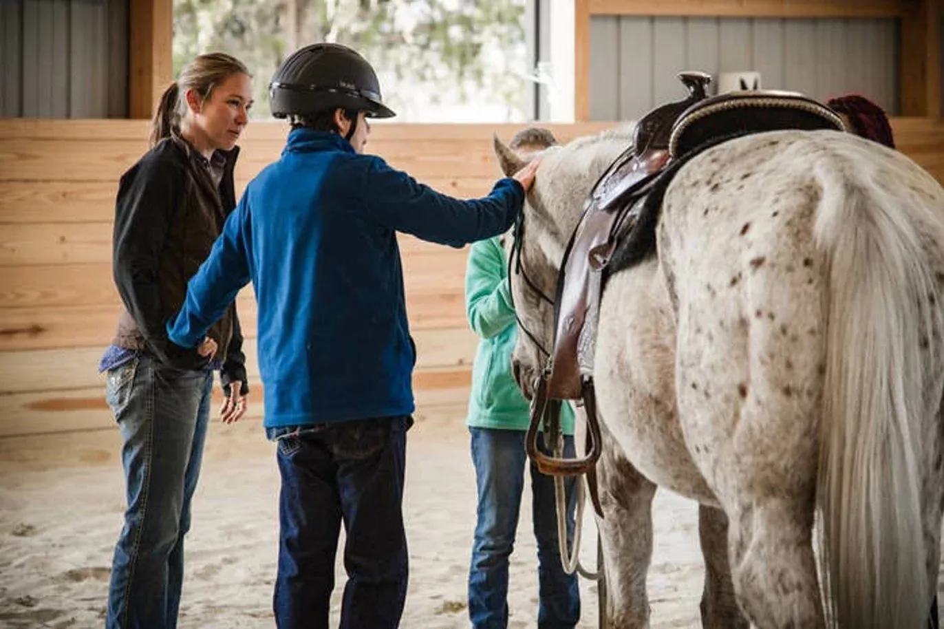 Un bambino con il casco accarezza un cavallo pezzato in un maneggio coperto, accanto a due ragazze